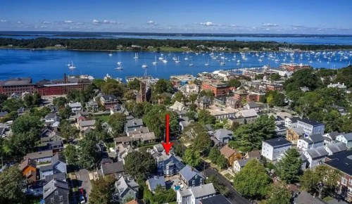 Aerial view of a coastal town with a harbor, boats, and a highlighted house among trees and buildings.