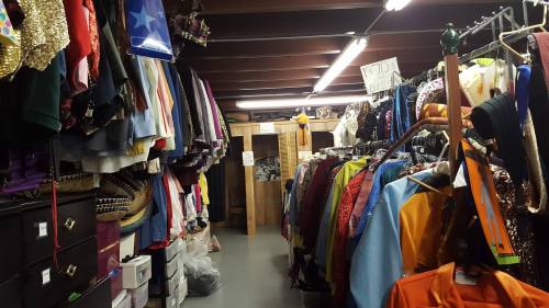 A cluttered costume storage area with colorful outfits hanging on racks and boxes stacked on the floor.