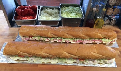 Two long sub sandwiches filled with layers of lettuce, tomatoes, and meats, placed on a wooden counter.