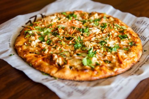 A freshly baked pizza topped with shredded chicken, carrots, and cilantro on a parchment-lined plate.