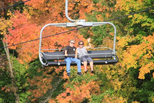 A couple smiles and waves while riding a chairlift surrounded by vibrant autumn foliage.