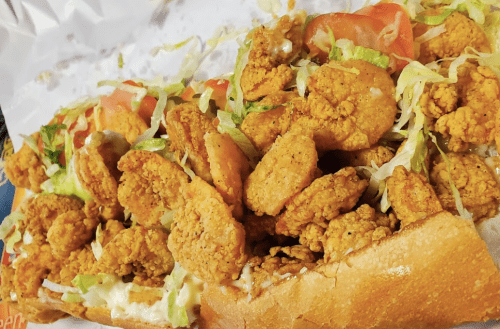 A close-up of a loaded sandwich filled with crispy fried shrimp, lettuce, and tomato on a toasted hoagie roll.