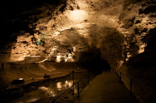 A dimly lit cave interior with rocky walls, a pathway, and a reflective water surface.