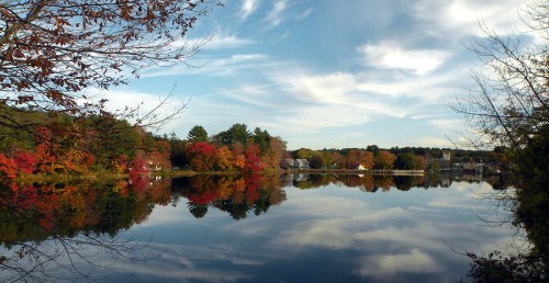 A serene lake reflecting colorful autumn trees and a cloudy sky, surrounded by lush greenery.