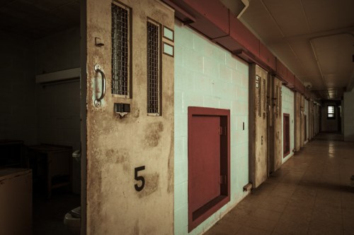 A dimly lit corridor in an old prison, featuring several closed cell doors with numbers and barred windows.