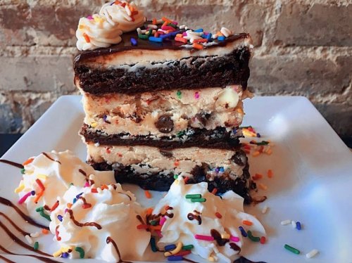 A tall slice of layered dessert with chocolate cake, cookie dough, and whipped cream, topped with colorful sprinkles.