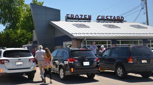 A busy frozen custard shop with customers walking in and parked cars outside on a sunny day.