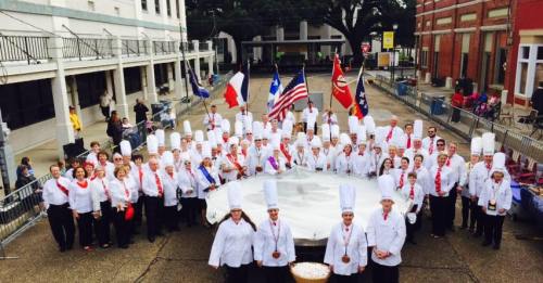 A large group of chefs in white uniforms and hats, holding flags, gathered around a giant cooking pot in a festive setting.