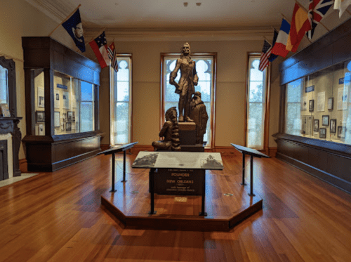 A museum interior featuring a statue of a historical figure, surrounded by flags and display cases with photographs.