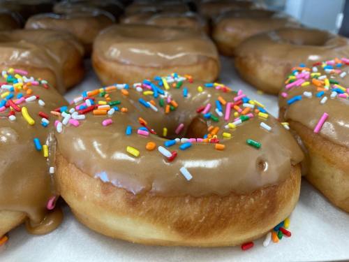 Close-up of a glazed donut topped with colorful sprinkles, surrounded by more donuts in a display case.