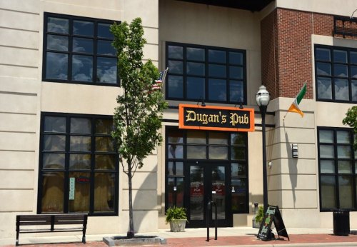 Exterior of Dugan's Pub featuring large windows, an orange sign, and a bench outside, with flags displayed.