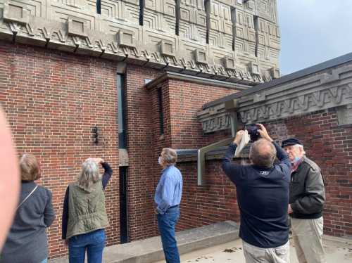 A group of people observes architectural details on a brick building, with one person taking a photo.