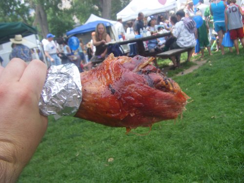 A hand holding a large, roasted turkey leg wrapped in foil, with a busy outdoor festival scene in the background.