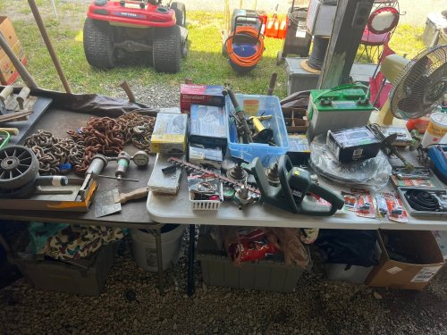 A cluttered table filled with various tools, hardware, and supplies, with a lawn tractor and storage in the background.