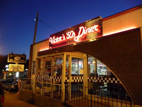 A retro diner with neon signage, featuring a checkered pattern and large glass windows, illuminated at dusk.