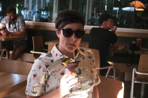 A person wearing sunglasses and a patterned shirt sips a drink in a cafe, with others in the background.