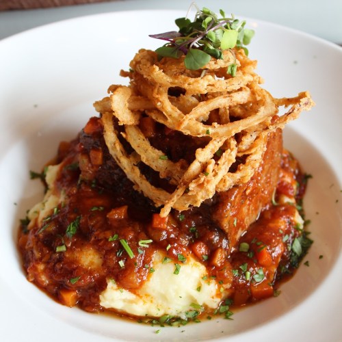 A plate of mashed potatoes topped with savory meat, rich sauce, and crispy onion rings, garnished with microgreens.