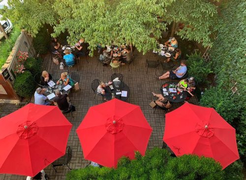 Aerial view of a bustling outdoor café with red umbrellas, surrounded by greenery and patrons enjoying their meals.