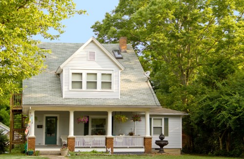 A charming two-story house with a green roof, white siding, and a porch surrounded by lush trees and plants.