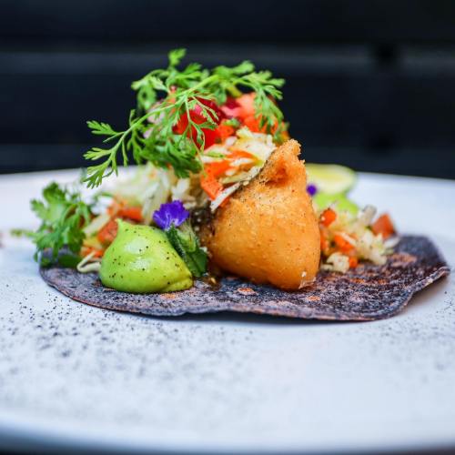 A vibrant taco on a blue corn tortilla, topped with slaw, avocado, and fresh herbs, garnished with edible flowers.