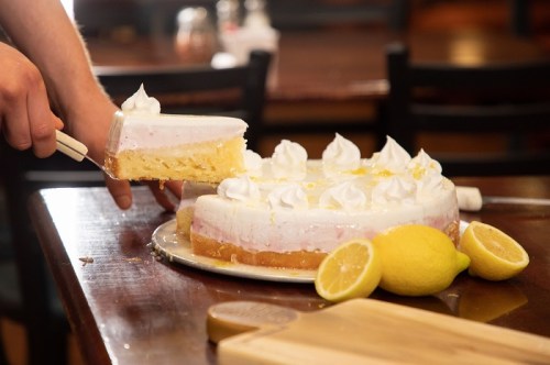 A person slices a lemon cake topped with whipped cream and lemon slices, with a wooden table in the background.