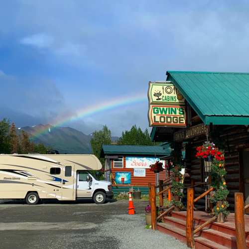A cozy lodge with a sign, an RV parked nearby, and a rainbow arching over a scenic mountain backdrop.