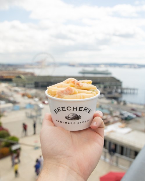 A hand holding a cup of mac and cheese from Beecher's, with a waterfront view in the background.