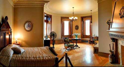 A vintage bedroom with a bed, fan, and seating area, featuring ornate decor and large windows.