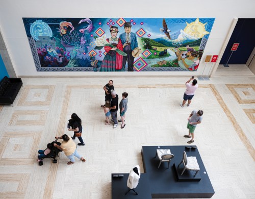 A vibrant mural on a wall in an art space, with people exploring the area below.