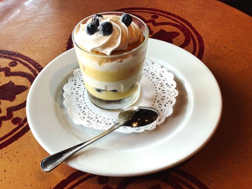 A layered dessert in a glass, topped with whipped cream and blueberries, served on a white plate with a spoon.