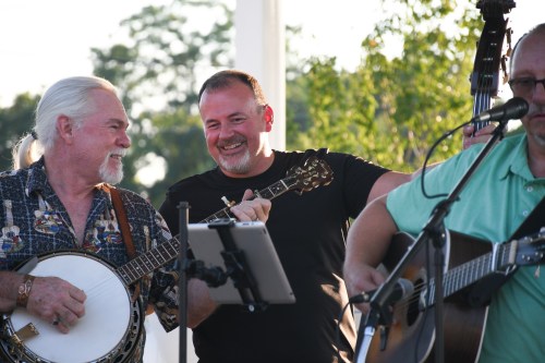 Three musicians perform outdoors, smiling and playing instruments, with greenery in the background.