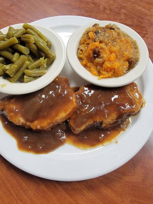 A plate with two pieces of meat covered in gravy, served with green beans and a bowl of sweet potato casserole.