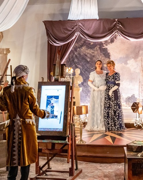A painter in period costume captures two women in elegant dresses against a dramatic backdrop in a gallery setting.