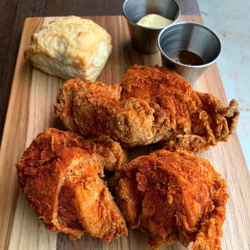 A wooden platter with three pieces of crispy fried chicken, a biscuit, and two dipping sauces in small cups.