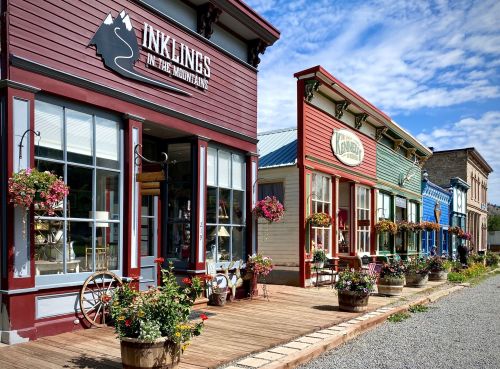 Colorful storefronts line a wooden walkway, adorned with flower pots and hanging baskets under a blue sky.