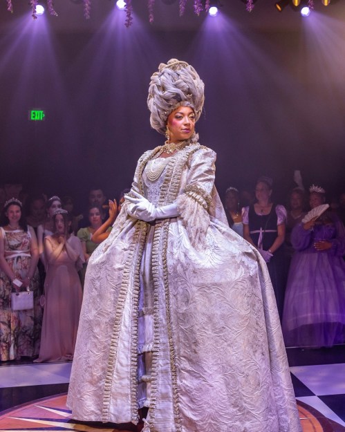 A performer in an elaborate silver gown and ornate hairstyle stands confidently on stage, surrounded by an audience.