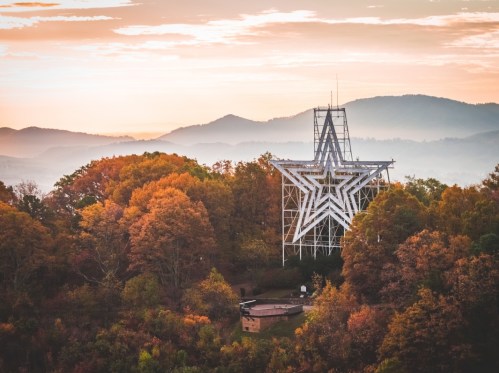 A large star-shaped structure stands amidst colorful autumn trees, with mountains and a soft sunrise in the background.