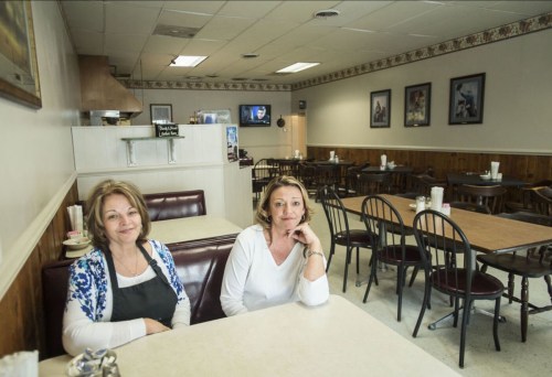 Two women sit at a table in a nearly empty diner, with a television and framed pictures on the walls in the background.