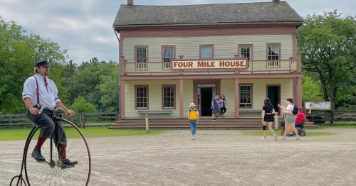 A vintage cyclist on a penny-farthing in front of the historic Four Mile House, with visitors exploring the area.