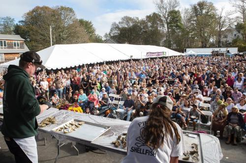 A large crowd watches a seafood event with chefs preparing oysters on stage under a tent.