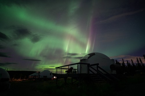 A night sky illuminated by vibrant green and purple auroras above dome-shaped structures in a serene landscape.