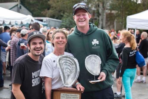 Three people smile while holding large oyster trophies at an outdoor event, with a crowd and tents in the background.