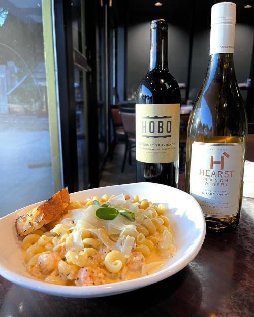 A bowl of pasta with shrimp, topped with cheese, alongside two bottles of wine on a table by a window.