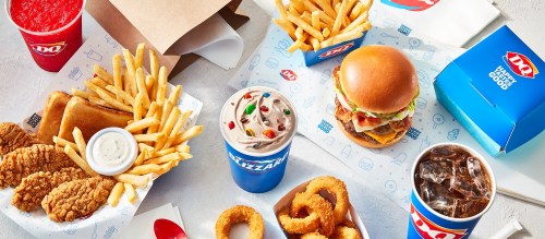 A spread of fast food including fries, chicken strips, a burger, a Blizzard, and drinks on a colorful table.