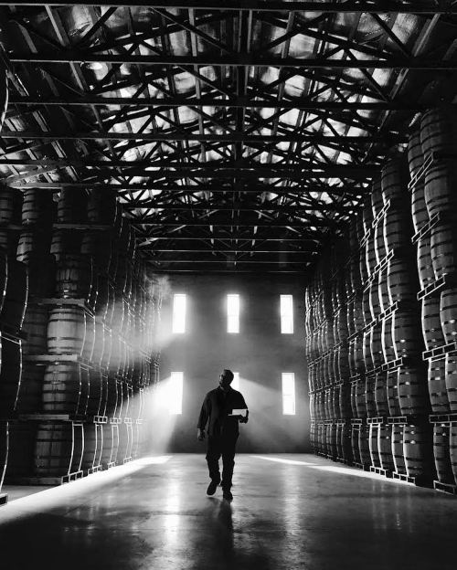 A silhouette of a person walking through a dimly lit warehouse filled with stacked barrels.
