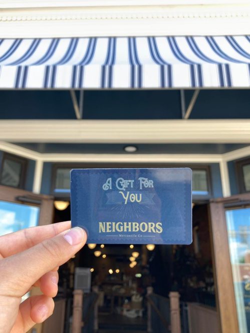 A hand holds a gift card labeled "A Gift For You" with "NEIGHBORS" printed below, in front of a storefront.