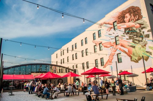 Outdoor gathering space with red umbrellas, people socializing, and a large mural of a woman on a building.