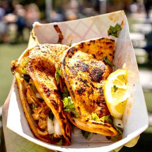 Three grilled tacos filled with meat and garnished with cilantro, served with a lemon wedge in a paper tray.