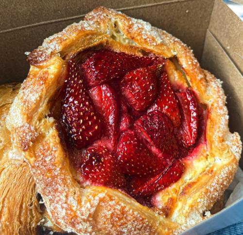A close-up of a pastry filled with fresh strawberries, topped with a sugary glaze, in a brown box.