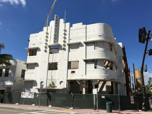 Art Deco building under renovation, featuring a white facade and construction equipment nearby, surrounded by green fencing.
